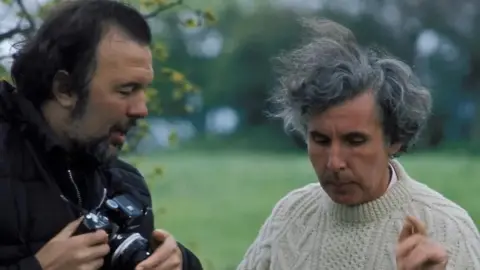 Rex Pyke Sir Peter Hall, a dark-haired, bearded man wearing a dark coat and holding a stills camera, looks at Ronald Blythe who is wearing a white jumping and pointing with the forefinger of his left hand