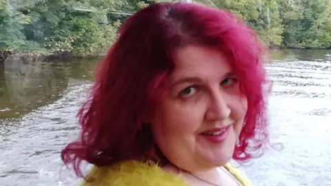 Handout A woman with shoulder-length bright red hair looks at the camera. She wears a yellow fluffy jumper and is standing in front of a window with trees and water in the background.