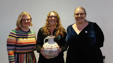 Syntax Poetry Collective Lizzy Dening, Lauren Kendrick and Hilary Steele standing in a row smiling into the camera. They are standing in front of a white wall. Lauren Kendrick, who is in the middle and has long, red hair, glasses and is wearing a brown top, is holding a large ceramic jug - her award for being named last year's Peterborough Poet Laureate.