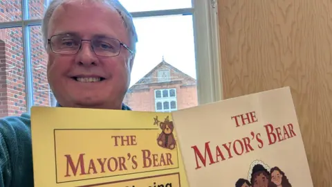 Jon Harvey Jon Harvey taking a selfie with his book and a sign inviting people to get their own books signed. He is smiling and wearing glasses.