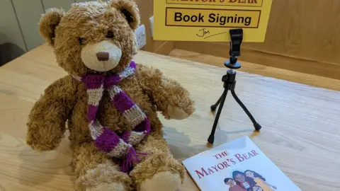 Jon Harvey A teddy bear is sat on a table wearing a purple and white scarf. Next to him is a copy of The Mayor's Bear book.