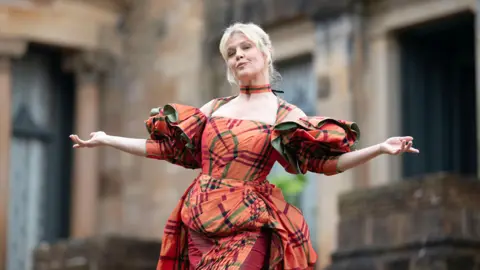 Mark Mainz A woman, Ashley Jensen, with blonde hair poses in front of a grand looking building. She has both her arms outstretched and is pouting. She is wearing a costume dress which is or ange and green tartan.