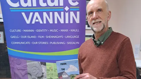 CULTURE VANNIN David Callin, who has grey hair and a grey beard and it wearing a red jumper. He is standing next to a banner that reads Culture Vannin. 