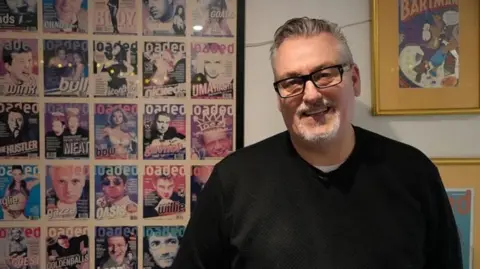 Tim Southwell standing in a room wearing black jumper and glasses in front of a montage of Loaded covers