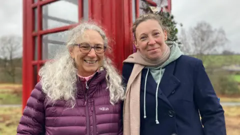 Kathy wearing a purple padded jacket and tortoiseshell glasses. She has long grey curly hair and is smiling at the camera. Standing next to her is Vicky, wearing a mint-green hoodie underneath a blue buttoned coat, and a pale pink scarf. She has blonde curly hair which is tied back in a bun. They are both standing in front of the red telephone box, which is in the middle of a field area.