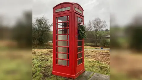 BBC A disused telephone box in the middle of a freshly cut field. There are grey slate slabs beside it, and cut grass strewn all over the ground. It is a wet overcast day and the sky is grey. The Perspex panels on the phone box are clouded and the panel at the top which reads 'Telephone' is barely legible. A Christmas wreath has  been placed on the door with a white bow on it.