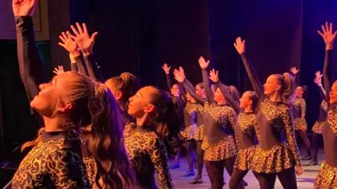 Sharon Higgins A side view of Bethan and others in cheetah-print dresses dancing with one arm extended above theme on a stage