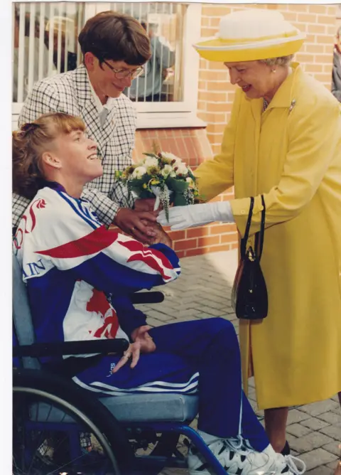 SUPPLIED A young woman in a tracksuit hands flowers to the Queen dressed in a yellow coat and white gloves