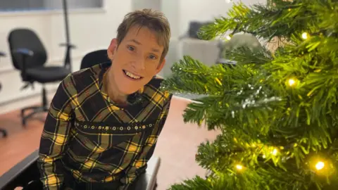 Simon Thake A woman in a yellow dress sits in a wheelchair smiling next to a sparkly Christmas tree