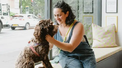 Colours of Arley Vicki Shenkin Kerr and her dog sit in her bookshop window. Ms Shenkin Kerr wears denim dungarees. She has dark, curly hair. Her dog has brown, curly fur.