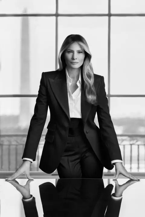 Regine Mahaux / The White House Melania Trump with her hands on the desk in a black and white suit with the Washington monument behind her