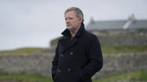 BBC/Silverprint Pictures/Mark Mainz Douglas Henshall in charcter as DI Jimmy Perez, he is wearing a black coat and is looking into the distance