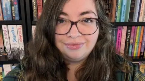 Bethan Hindmarch Bethan Hindmarch who has long dark hair and glasses and is wearing a green check shirt, in front of a bookcase full of books