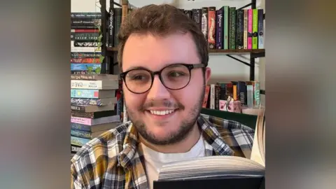 Scholastic Bill Wood has brown hair and brown facial hair. He is wearing black glasses and a yellow, white and black shirt. He is holding a black book that is open in front of him.