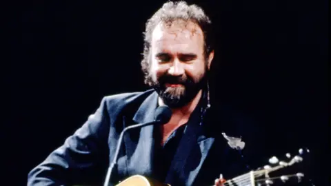 John Martyn performing onstage - he has an acoustic guitar and is wearing a dark suit.