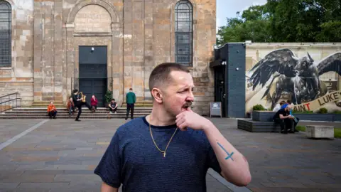 Blue Rose Code Ross Wilson is standing at the forefront of the picture, with a dark T-shirt on and a gold cross hanging around his neck. He is looking to the right of the picture. Behind him is a converted church, where members of his band are standing or sitting on the steps