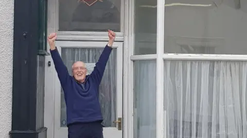 Dorothy Lambert Charlie Lambert celebrating outside Norman Nicholson's  house after the property was bought in February. Mr Lambert is smiling and his hands are up in the air. He is wearing a navy blue sweatshirt and has glasses. The house has large front windows with white panes. 