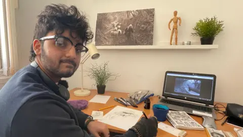 BBC Aaban sits at a desk. On the wall is a photograph in black and white taken by the photographer James Ravillious of a woman herding sheep. On the desk he has ink, paper and sketches for his graphic novel, Widening Gyre.