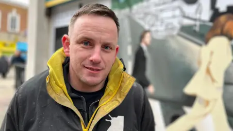 Emma Baugh/BBC Nathan Murdoch, a man who is looking at the camera and smiling. He is wearing a yellow and black jacket and is stood in front of a mural he has been creating. 