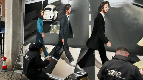 Emma Baugh/BBC Two men in black overalls are crouched by the floor working on the mural.