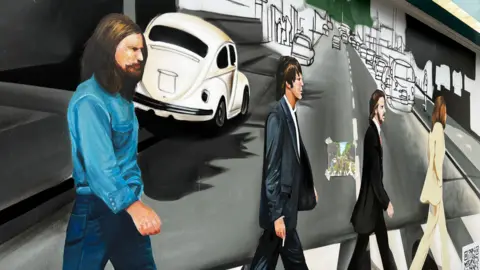 Emma Baugh/BBC A mural depicting the four band members of The Beatles walking over a zebra crossing. One is in a white suit, two are in dark suits and the fourth is a denim top and jeans. Cars are depicted in the background. 