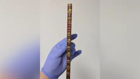 Moyse's Hall Museum A view of the book's thin binding. It is brown with some gold details. A person wearing blue gloves holds it.