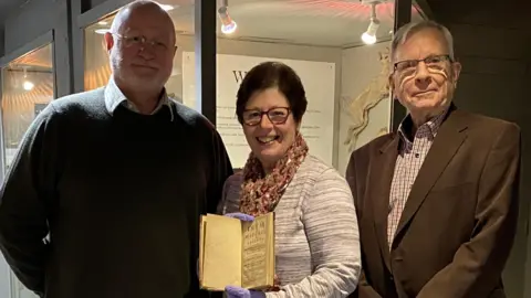 West Suffolk Council Two men stand alongside a woman who holds the book while she wears blue gloves. They smile at the camera.