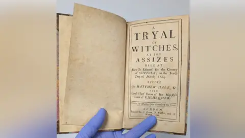 Moyse's Hall Museum The historic book that recounts the Bury St Edmunds witch trials. It shows the first page of the book, which someone wearing blue gloves holds open.