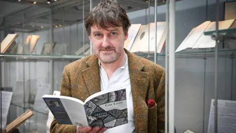 PA Media Peter Mackay is standing near display cases containing rare books. He is holding a copy of one his books of poetry.