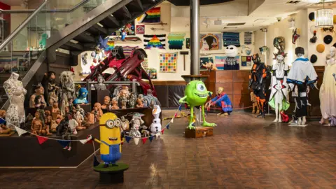 Thierrry Bal A room full of various items donated by hobbyists. There is a large yellow Minion figurine and a large Mike Wazowski figure from Monsters Inc, which is bright green. Behind them are costumes on mannequins and various small sculptures of busts or people.