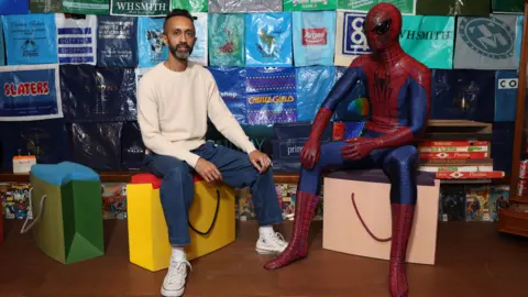 Lia Toby Hetain Patel is sitting on a stool that looks like a yellow shopping bag. He has short dark hair and dark and grey facial hair. Sitting next to him on a similar stool is a life-size model of Spider-Man in a blue and red suit. In the background is a wall made up of branded plastic bags.