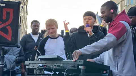 BBC Three men stand in front of a DJ mixer with people gathered behind them. Ed Shearan, on the left, has short ginger hair and is wearing a white t-shirt with black trim markings. Next to him is a young man with a microphone in his left hand and on the far right is a man wearing a grey and red hoody with his hands on the mixing table.