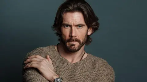 Martin Sylvester A headshot of James Wilkinson in a brown knitted jumper. He is posing while sat down in front of a dark green background. He has long brown hair and a dark beard.