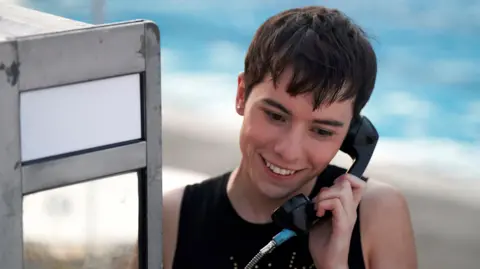 BBC/Hera/Enda Bowe Ellis Howard as Byron in What It Feels Like For A Girl, using a pay phone and smiling.