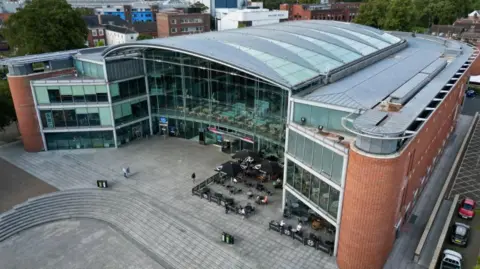 Martin Barber/BBC A drone image of The Forum building. There is a glass atrium and a curved main roof, with flatter roofs around the sides of the U-shaped building. The BBC office is in the left-hand wing. There is a paved area, stone steps as well as tables and chairs outside the building.