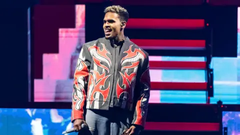Getty Images Chris Brown, who will perform at Cardiff's Principality Stadium on Thursday, pictured performing in Detroit Michigan in April. He wears a black jacket with red flames on it, with a microphone in his left hand. He looks away from the camera and pokes his tongue out to the side of his teeth.