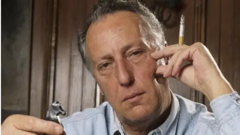 Getty Images Frederick Forsyth is looking at the camera without smiling. He is holding a cigarette and lighter. He looks in his 40s or 50s.