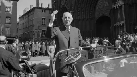 Getty Images General de Gaulle is seen in a black and white photo in civilian dress. He is standing in the back of an open top car waving at the crowd.