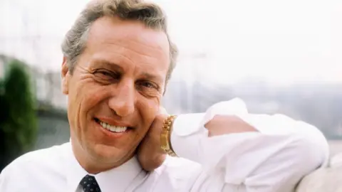 Getty Images Frederick Forsyth photographed in the 1980s. He is smiling at the camera and his his hand on his neck.