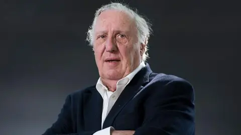 Getty Images Frederick Forsyth is seen in later life. He is looking slightly off camera in a head and shoulders portrait. His arms are folded and he is wearing a white shirt and blue jacket.