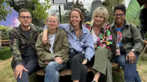 BBC Five people sitting on a wooden chair. Alison Howe has short light-coloured hair and glasses, wearing a green anorak and white T-shirt. Emily Eavis has long brown hair and is wearing a blue and white shirt. There are potted plants behind them.