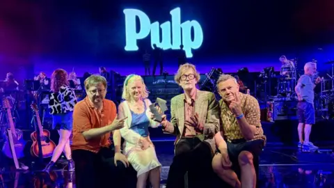 Official Charts The band Pulp are sat on a stage holding their award from the Official Charts
