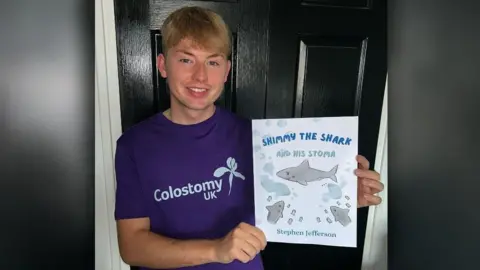 Supplied Stephen Jefferson holding a copy of his book Shimmy the shark and his stoma. The book cover is white, with a blue title and several sharks. Mr Jefferson is smiling at the camera and wearing a purple Colostomy UK t-shirt. He has blond hair and blue eyes.