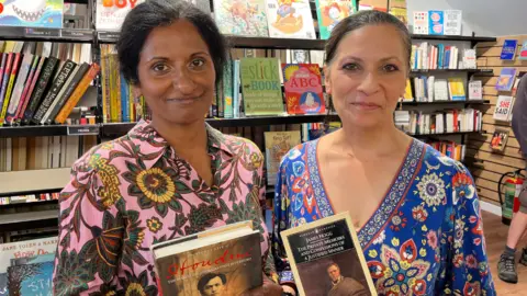 Juliette Parkin/BBC Sisters Seeta and Lizzie Seetharaman pictured with their books at the Oxfam bookshop in Hove, East Sussex. Seeta bought a biography of Houdini while Lizzie bought The Private Memoirs and Confessions of a Justified Sinner. Seeta is wearing a pink and green floral dress, while Lizzie is wearing a blue, red and yellow dress.