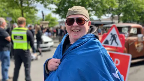 BBC Andy Edwards, a man wearing sunglases, is draped in a blue flag. He is smiling for the camera.