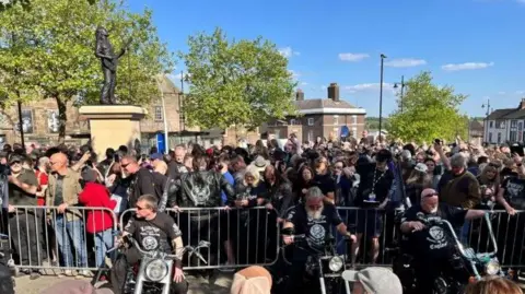 Three men on bikes in front of a railing, behind which is a crowd of people next to the new statue of Lemmy.