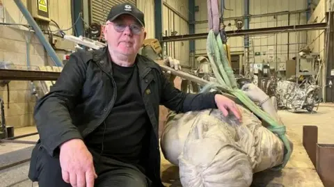BBC A man with a black hat, glasses, a black jacket, black shirt and jeans. He is sat down on a work table in a workshop and has his left hand on a statue of Lemmy, which is lying down.