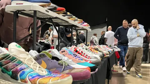 BBC A display table at a sneaker convention showcasing a variety of colourful and unique sneakers. The foreground features vibrant designs, including iridescent, multicolour, and patterned styles, arranged neatly in rows on black tablecloths. Additional sneakers are placed on an elevated rack above the main table. In the background, several people are standing and browsing other tables in a spacious indoor venue with polished concrete floors and dark walls.