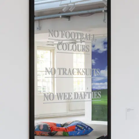Trackie McLeod A mirror has lettering on it reading "no football colours, no tracksuits, no wee dafties". In  the mirror you can see a reflection if his wider exhibition. 