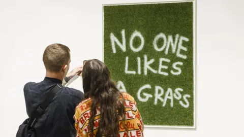 Trackie McLeod An image of green grass hangs on a gallery wall, spray painted on it in white, is the phrase "no one likes a grass". A man and a woman stand in front of the piece, taking a picture. 
